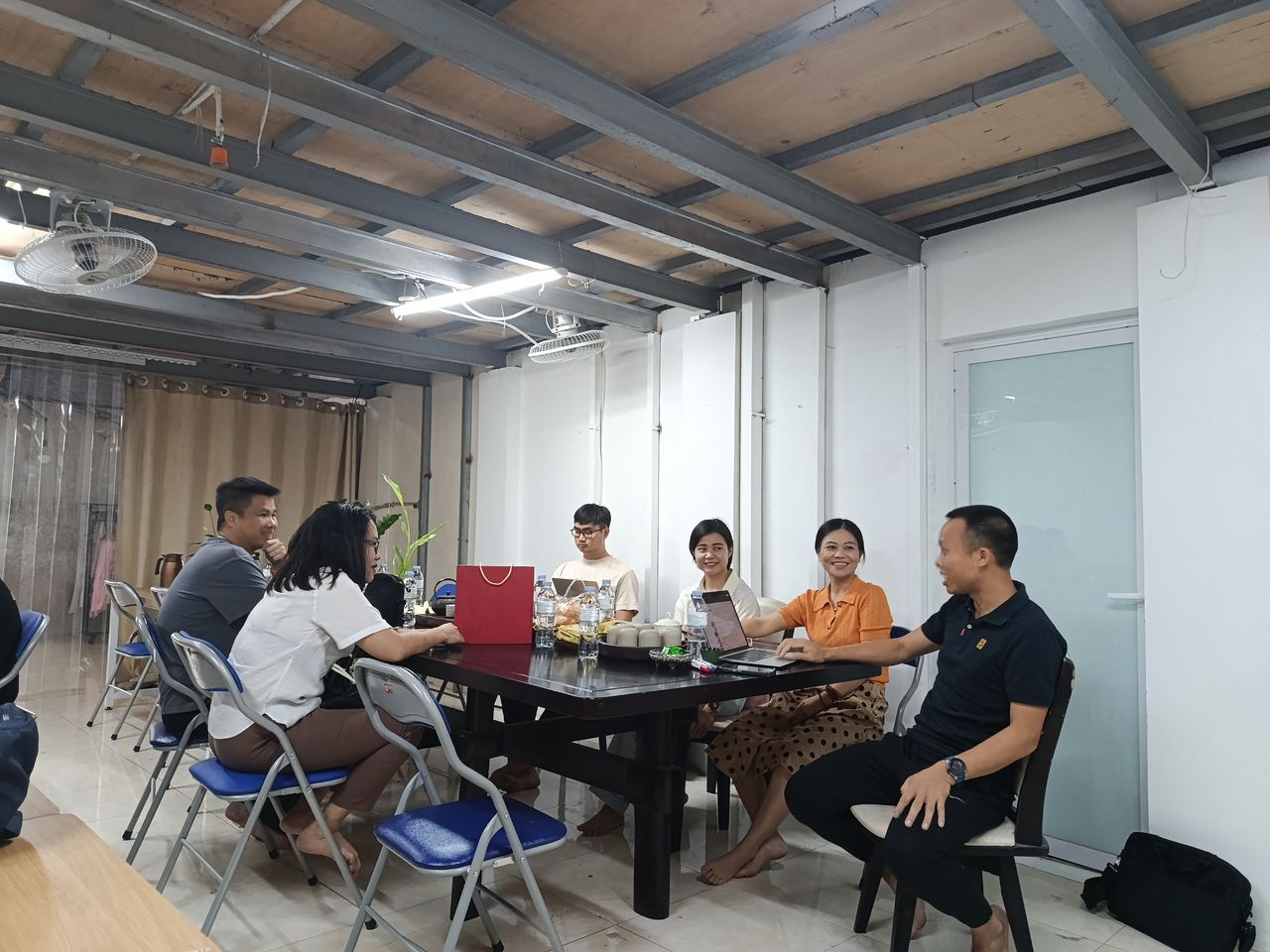 Sáu bảy người quanh bàn, laptop mở, túi quà đỏ